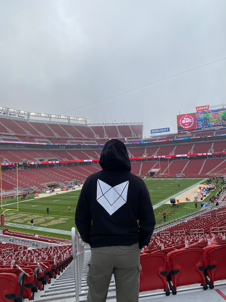 A man in a Cool Cat sweatshirt stands before the field of the San Francisco 49ers/Levi’s Stadium.