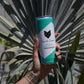 A hand holds a can of Cool Cat Elderflower Sparkling Cocktail in front of a palm tree.