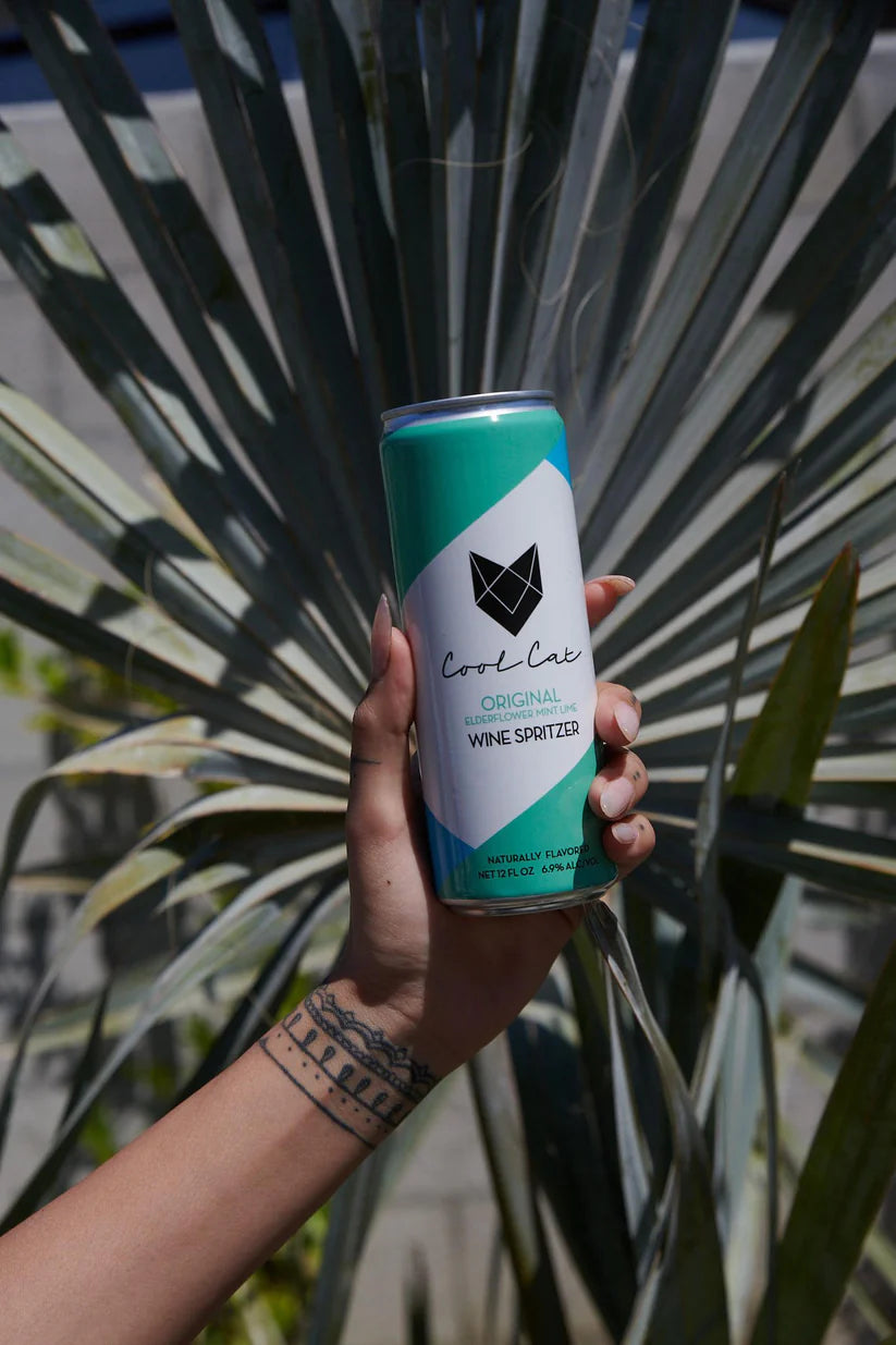 A hand holds a can of Cool Cat Elderflower Sparkling Cocktail in front of a palm tree.